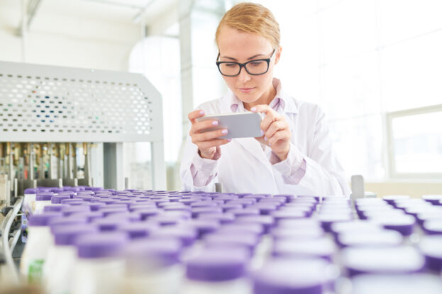 Beautiful inspector wearing eyeglasses and white coat taking picture on modern smartphone while checking quality of soy milk at production department of dairy plant